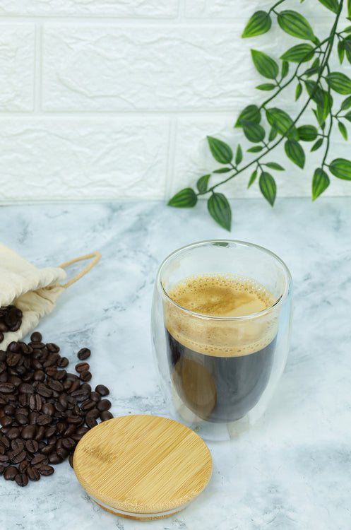 Glass of coffee with a wooden lid on a marble surface with coffee beans and a plant in the background.
