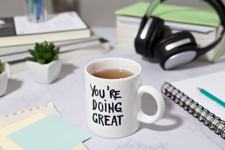 Mug with 'You're Doing Great' text on a desk with books and headphones