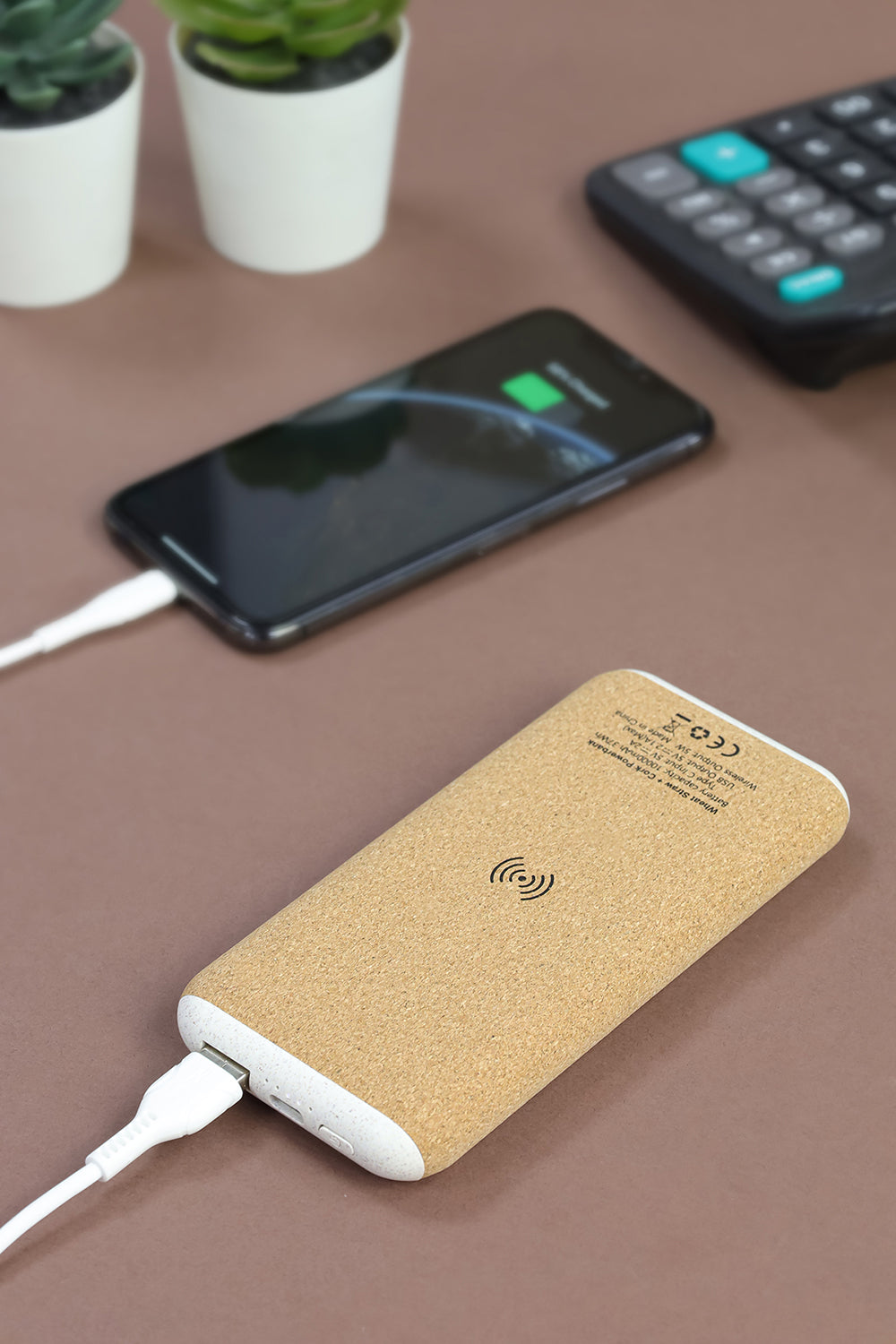 A beige power bank with cork finish being used to charge a smartphone, placed on a brown surface with a potted plant and a calculator in the background.