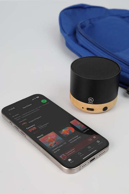 Black portable speaker next to a smartphone on a light gray surface with a blue bag in the background.