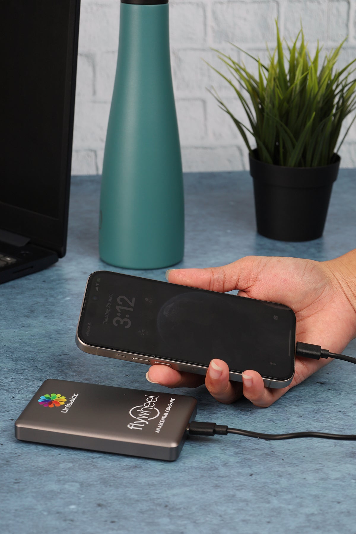 Person holding a smartphone connected to a portable power bank on a table with a teal bottle and plant in the background.