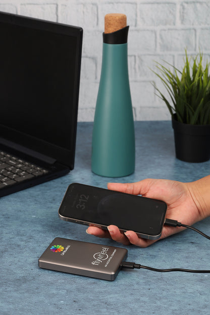 Person holding a smartphone with a wireless charging pad on a desk with a laptop, water bottle, and plant.