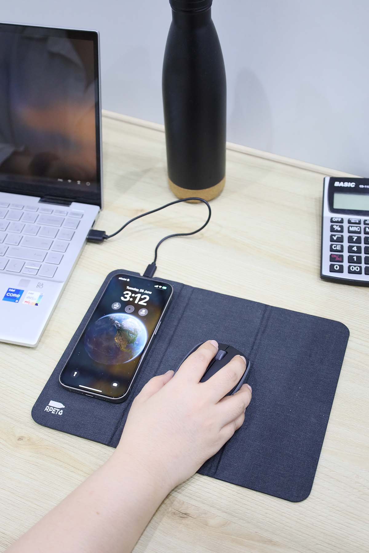 Hand using a mouse on a desk with a laptop, phone, and water bottle in the background.