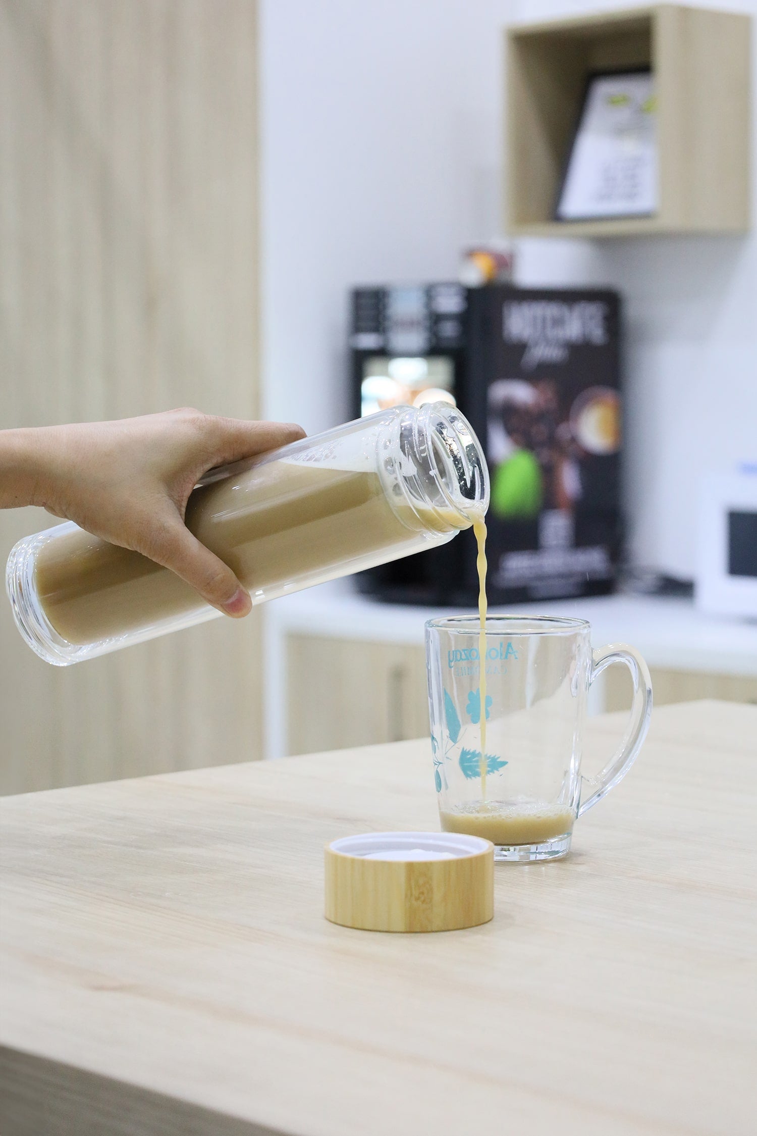 Person pouring a liquid from a glass container into a mug on a wooden surface.