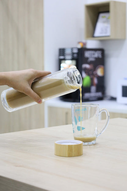 Person pouring a liquid from a glass container into a mug on a wooden surface.