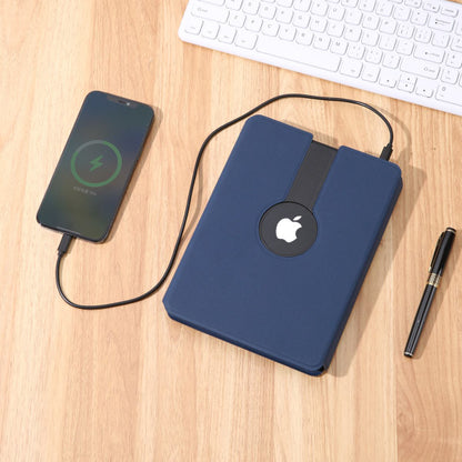Blue notebook with Apple logo, smartphone charging, and keyboard on a wooden surface